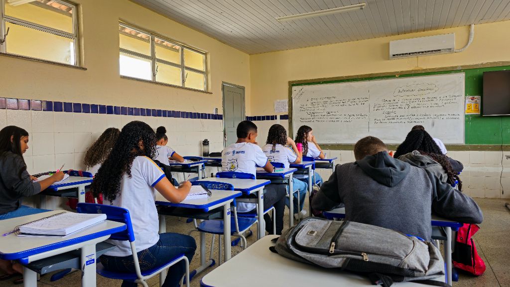 Alunos em sala de aula do ensino público escrevem em cadernos durante atividade escolar, com quadro branco ao fundo com anotações. A imagem representa o contexto do Fundeb e o financiamento escolar na educação básica brasileira.