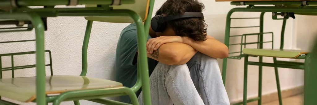 Estudante sentado no chão de uma sala de aula, encostado à parede e entre carteiras verdes, com fones de ouvido e a cabeça apoiada nos braços, em postura que sugere isolamento ou tristeza.