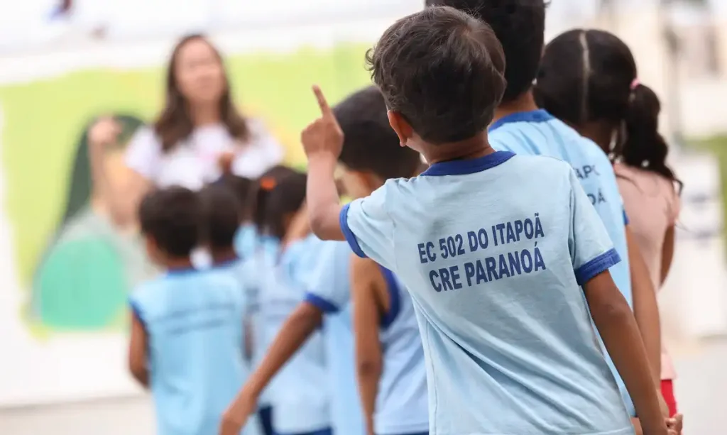 Crianças pequenas vestindo uniformes azul-claro caminham em fila dentro de uma escola, enquanto uma professora aparece desfocada ao fundo. Em destaque, um menino levanta o dedo e usa camiseta com a inscrição “EC 502 do Itapoã – CRE Paranoá”.