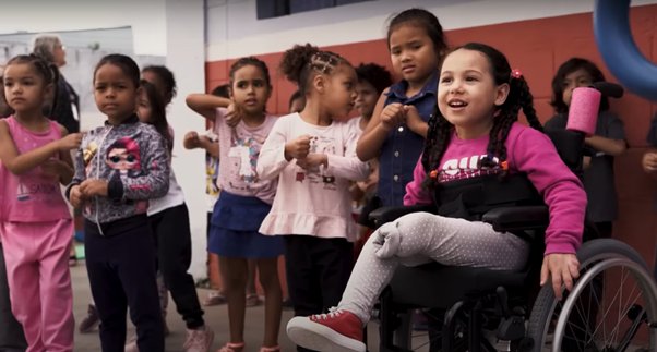 Menina cadeirante participa de atividade escolar com outras crianças em sala de aula, representando educação inclusiva, inclusão escolar e uso de cadeira de rodas.
