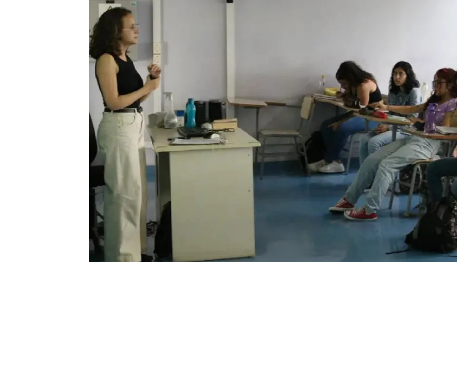 Professora em pé diante da turma conduz aula em sala iluminada, enquanto estudantes sentados escutam e fazem anotações.