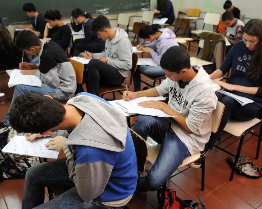 Estudantes sentados em carteiras realizam prova em sala de aula. Jovens estão concentrados preenchendo o caderno de questões e folhas de resposta, representando o momento do Enem.
