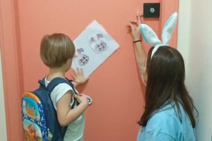 Criança com mochila olha desenho de coelhinhos na porta enquanto adulta com tiara de orelhas de coelho aponta para o papel.